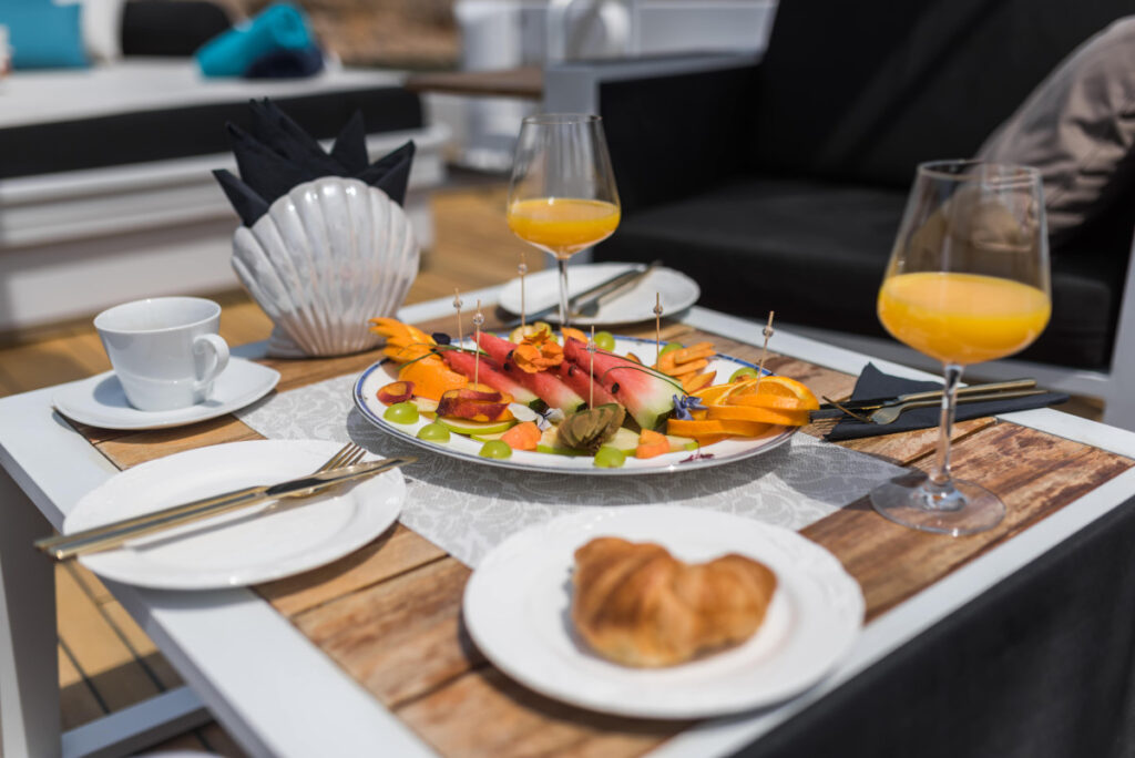 A gourmet breakfast spread on a yacht deck featuring fresh espresso, pastries, and tropical fruit with a background view of a quiet, sun-drenched coastal cove.