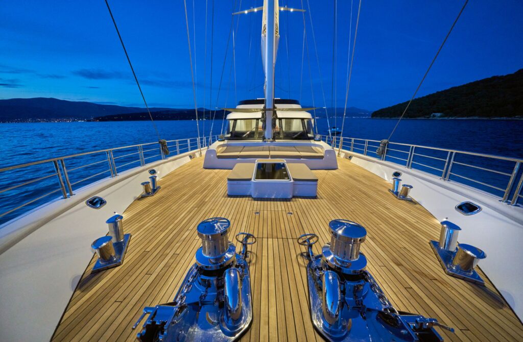 The teak deck of a luxury yacht at dusk, overlooking a calm sea and distant island silhouettes under a soft purple and orange sunset sky.