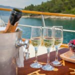 A polished stainless steel ice bucket containing a bottle of rosé and a bottle of champagne, sitting on a high-gloss teak wood table alongside four elegant crystal wine glasses on the outdoor deck of a luxury yacht at sunset.