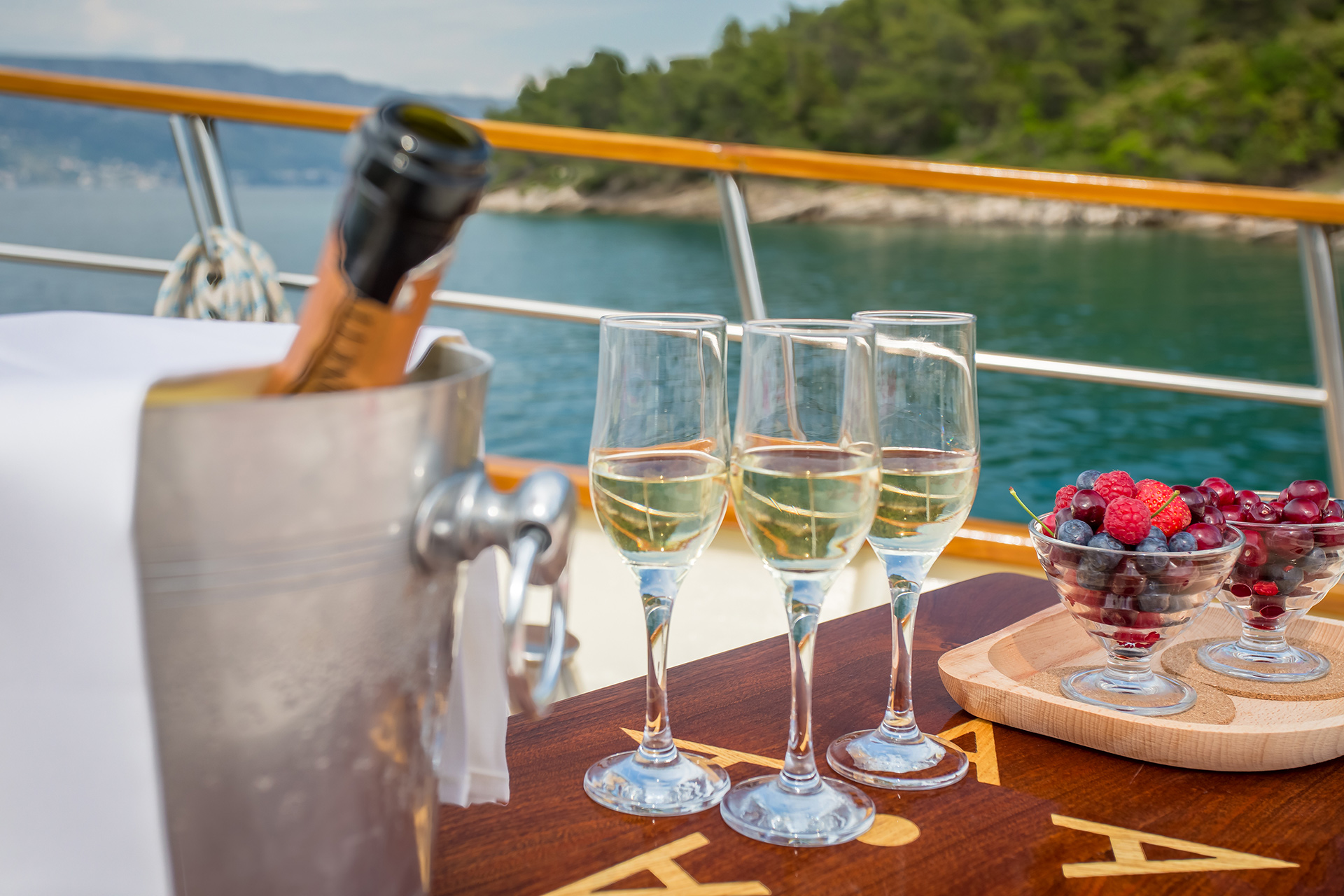 A polished stainless steel ice bucket containing a bottle of rosé and a bottle of champagne, sitting on a high-gloss teak wood table alongside four elegant crystal wine glasses on the outdoor deck of a luxury yacht at sunset.
