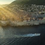 A luxury sailing yacht gliding past the ancient stone walls of Dubrovnik, Croatia during a golden sunset.