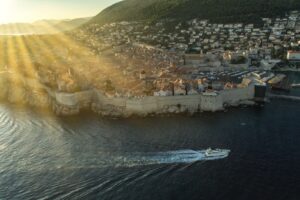A luxury sailing yacht gliding past the ancient stone walls of Dubrovnik, Croatia during a golden sunset.
